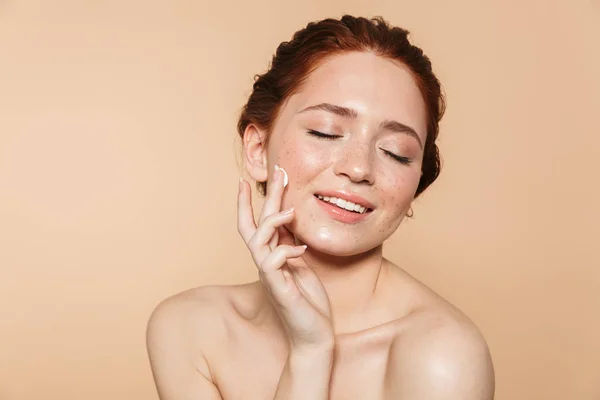 Incredibile giovane rossa donna posa isolato su sfondo parete beige prendersi cura della sua pelle con crema . — Foto Stock