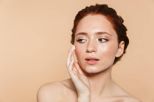Amazing young redhead woman posing isolated over beige wall background take care of her skin with cream.