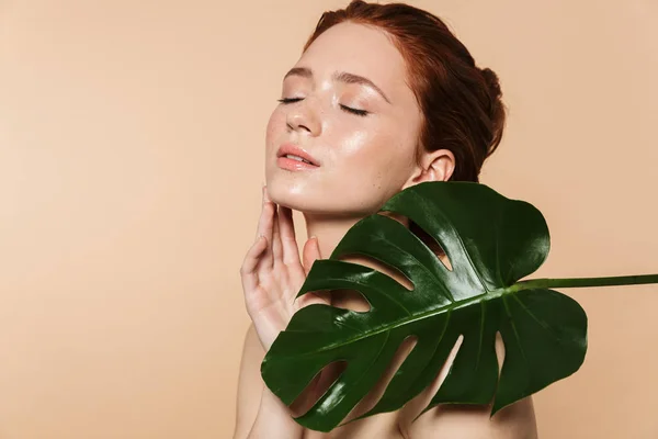 Mooie jonge roodharige vrouw poseren geïsoleerd over beige muur achtergrond met blad groene bloemen. — Stockfoto