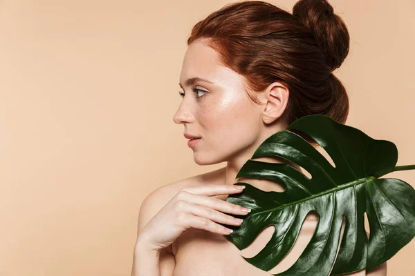 Mooie jonge roodharige vrouw poseren geïsoleerd over beige muur achtergrond met blad groene bloemen. — Stockfoto