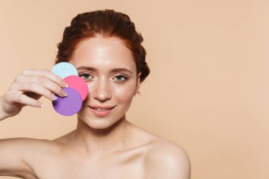 İnanılmaz genç kızıl saçlı kadın makyaj süngerleri tutarak bej duvar arka plan üzerinde izole poz