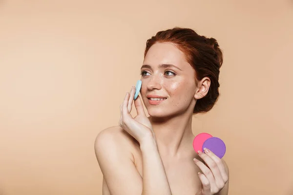 Verbazingwekkende jonge roodharige vrouw poseren geïsoleerd over beige muur achtergrond Holding make-up sponzen — Stockfoto