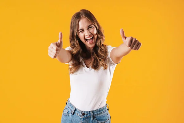 Menina sorridente emocional feliz em casual t-shirt branca posando isolado sobre fundo de parede amarela mostrando polegares para cima . — Fotografia de Stock