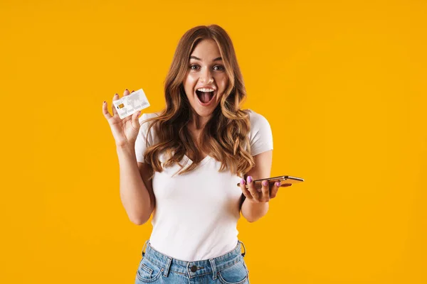 Foto de mulher muito jovem se alegrando enquanto segurando cartão de crédito e smartphone isolado sobre fundo amarelo — Fotografia de Stock