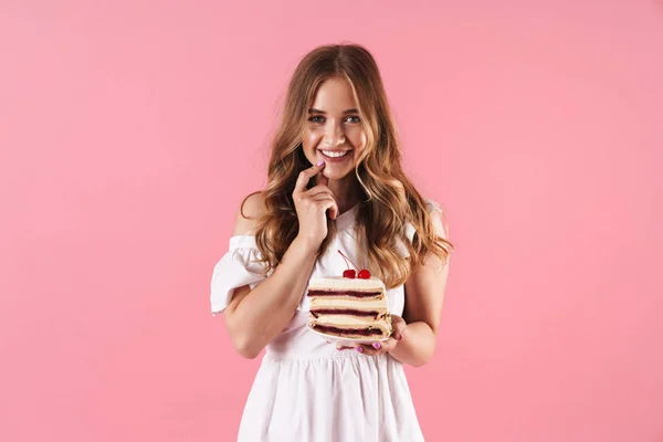 Bild av lycklig leende kvinna klädd i vit klänning tittar på kameran med fingret på hakan och håller bit av kakan — Stockfoto