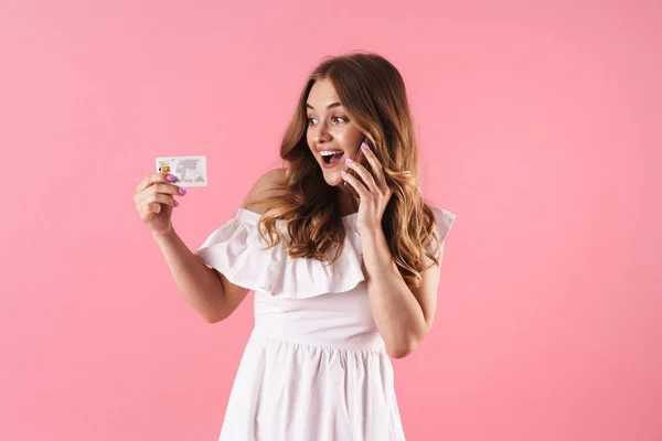 Boldog vidám fiatal csinos nő jelentő elszigetelt mint rózsaszín fal háttérben beszél mobiltelefon Holding hitelkártya. — Stock Fotó