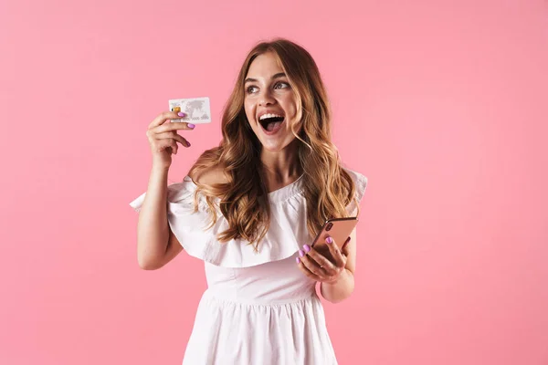 Glad glad ung söt kvinna poserar isolerade över rosa vägg bakgrund med hjälp av mobiltelefon Holding kreditkort. — Stockfoto