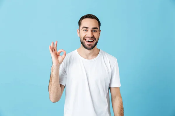 Sorrindo jovem homem barbudo feliz posando isolado sobre fundo parede azul mostrando gesto ok. Conceito de verão . — Fotografia de Stock