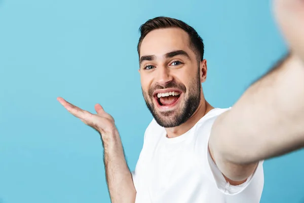 Sonriente joven hombre barbudo feliz posando aislado sobre fondo de pared azul tomar una selfie por cámara . — Foto de Stock
