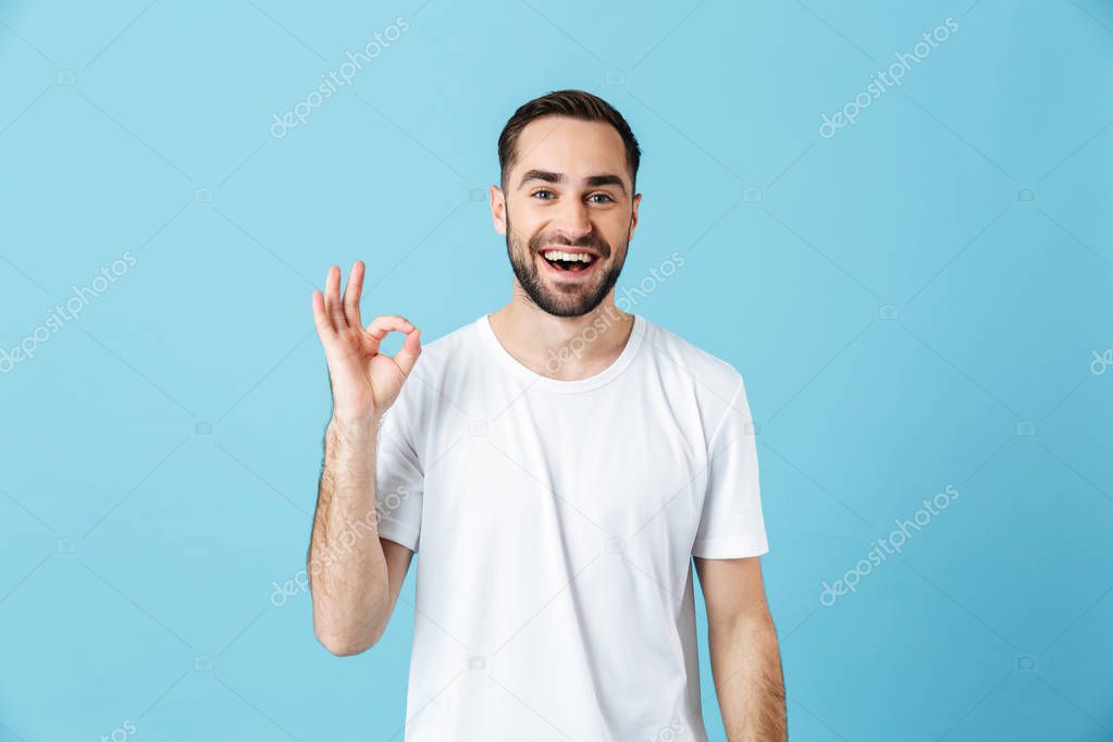 Smiling young happy bearded man posing isolated over blue wall background showing okay gesture. Summer concept.