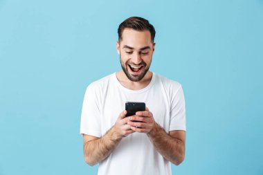 Heyecanlı genç mutlu sakallı adam cep telefonu sohbet kullanarak mavi duvar arka plan üzerinde izole poz.