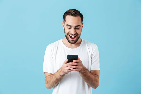 Izgatott fiatal boldog szakállas férfi pózol elszigetelt kék fal háttér segítségével mobiltelefon csevegés. — Stock Fotó