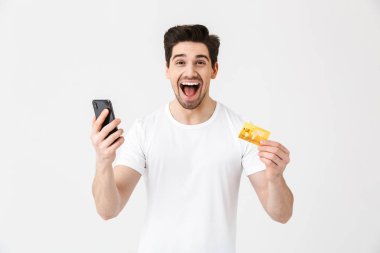 Heyecanlı mutlu genç adam kredi kartı tutan cep telefonu kullanarak beyaz duvar arka plan üzerinde izole poz.