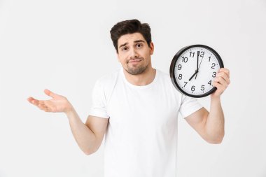 Şaşkın genç adam saat tutan beyaz duvar arka plan üzerinde izole poz.
