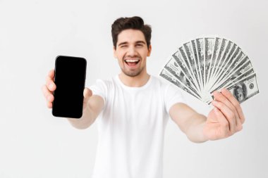 Heyecanlı mutlu genç adam cep telefonu ekran gösteren para tutarak beyaz duvar arka plan üzerinde izole poz.