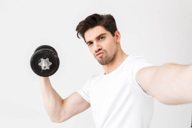 Ciddi genç adam beyaz duvar üzerinde dumbbell tutarak izole poz egzersiz yapmak kamera ile selfie çekmek.