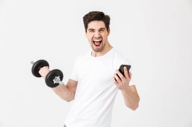 Duygusal mutlu heyecanlı genç adam cep telefonu kullanarak egzersiz yapmak dumbbell tutarak beyaz duvar üzerinde izole poz.