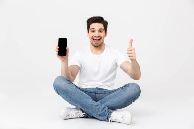 Heyecanlı genç adam ekranda oturan gösteren gösteren cep telefonu kullanarak beyaz duvar arka plan üzerinde izole poz.