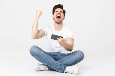 Heyecanlı genç adam cep telefonu ile beyaz duvar arka plan üzerinde izole poz kazanan jest yapmak oyunlar.