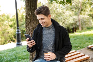 Cep telefonu kahve içme kullanarak yeşil parkta açık havada yürüyüş rahat giyim yakışıklı neşeli genç mutlu adam.