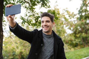 Yeşil parkta cep telefonu kullanarak açık havada yürüyen rahat giyimli genç adam selfie çekmek.