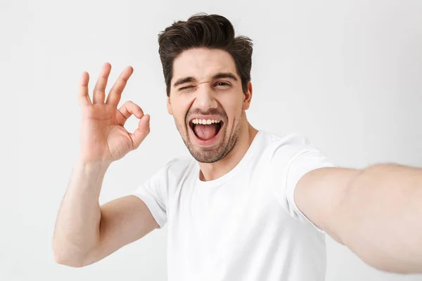 Aufgeregter glücklicher junger Mann posiert isoliert über weißem Wandhintergrund und macht ein Selfie mit der Kamera. — Stockfoto