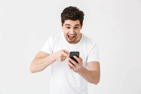 Upphetsad lycklig ung man poserar isolerade över vit vägg bakgrund med hjälp av mobiltelefon. — Stockfoto