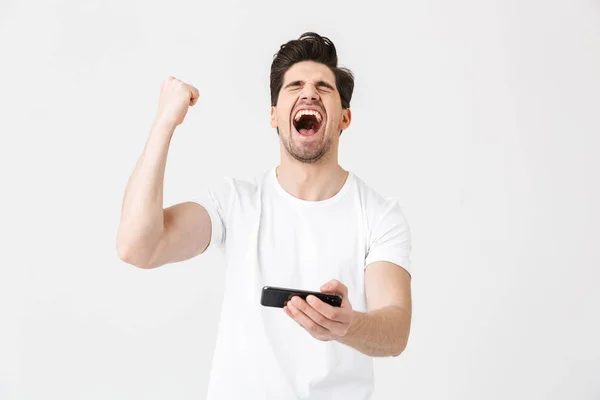 Izgatott boldog érzelmi fiatalember jelentő elszigetelt felett fehér fal háttérben játszani a mobiltelefonnal, hogy győztes gesztus. — Stock Fotó