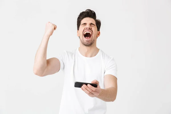 Emocionado feliz jovem emocional posando isolado sobre fundo da parede branca jogar jogos por telefone móvel fazer gesto vencedor . — Fotografia de Stock