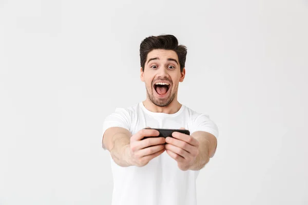 白い壁の背景の上に孤立したポーズ興奮幸せな感情的な若者は、携帯電話でゲームをプレイ. — ストック写真