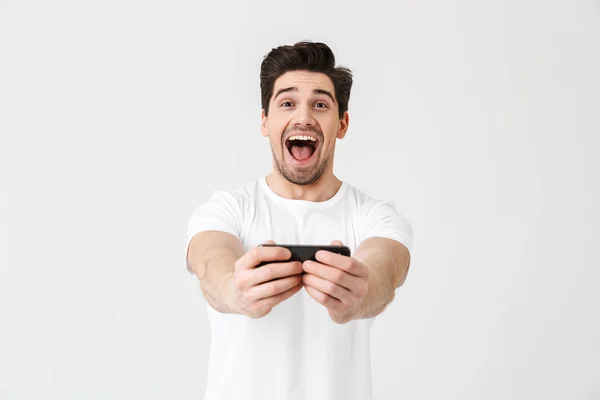 Opgewonden gelukkig emotionele jonge man poseren geïsoleerd over witte muur achtergrond spelen games door mobiele telefoon. — Stockfoto
