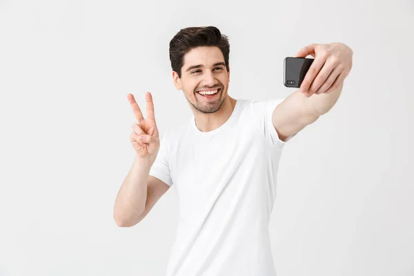 Emocional joven posando aislado sobre fondo blanco de la pared mostrando gesto de paz tomar selfie por teléfono móvil . — Foto de Stock