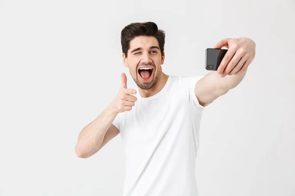 stock image Emotional young man posing isolated over white wall background pointing take selfie by mobile phone.