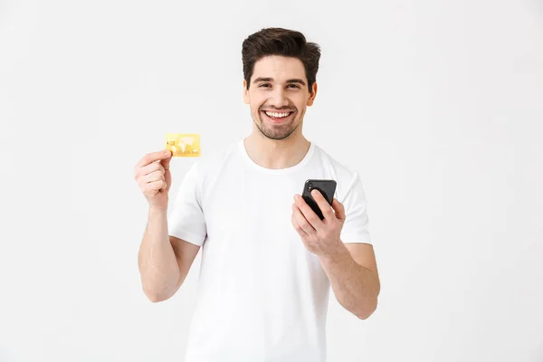 Emocionado joven feliz posando aislado sobre fondo blanco de la pared utilizando el teléfono móvil celebración de la tarjeta de crédito . — Foto de Stock