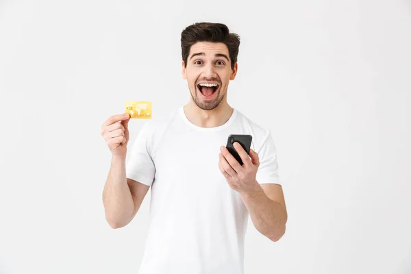 Upphetsad lycklig ung man poserar isolerade över vit vägg bakgrund med hjälp av mobiltelefon Holding kreditkort. — Stockfoto