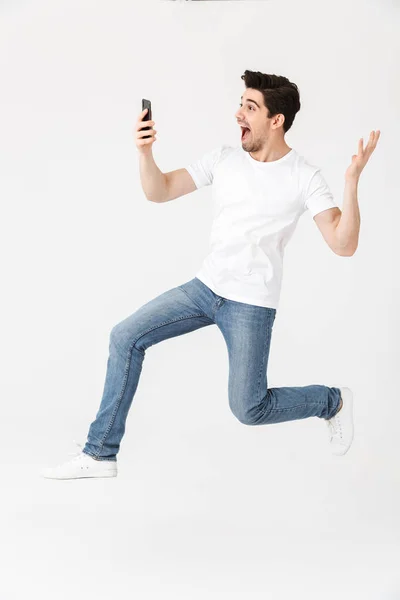 Happy young man posing isolated over white wall background using mobile phone jumping. — Stock Photo, Image