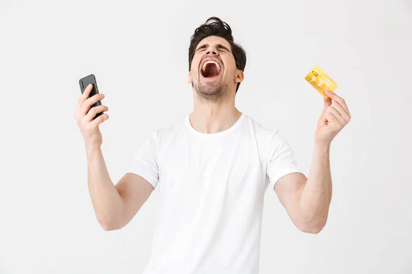 Opgewonden gelukkig jonge man poseren geïsoleerd over witte muur achtergrond met behulp van mobiele telefoon Holding creditcard. — Stockfoto