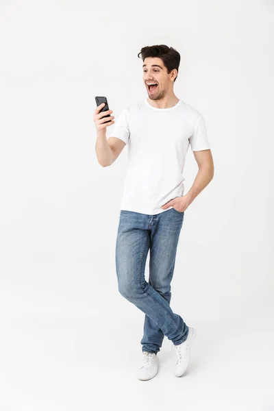 Jeune homme heureux excité posant isolé sur fond de mur blanc en utilisant le téléphone mobile . — Photo