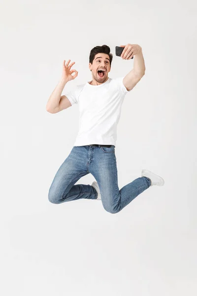 Young man posing isolated over white wall background using mobile phone take a selfie. — Stock Photo, Image