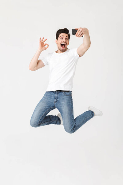 Young man posing isolated over white wall background using mobile phone take a selfie.