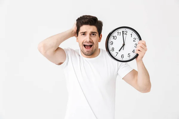 Urlando confuso giovane uomo posa isolato su sfondo bianco muro che tiene orologio . — Foto Stock