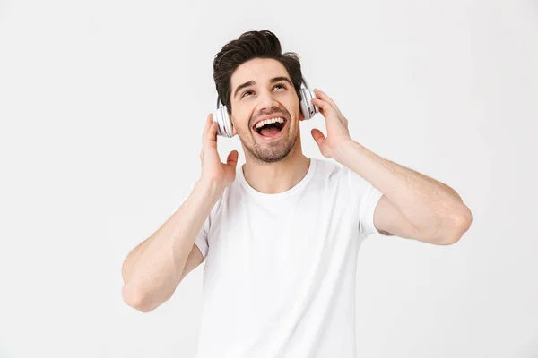 Emotioneel opgewonden jonge man poseren geïsoleerd over witte muur achtergrond luisteren muziek met koptelefoon. — Stockfoto