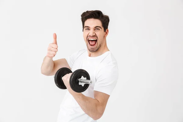 Emotional happy excited young man posing isolated over white wall holding dumbbell make exercise make thumbs up gesture. — Stock Photo, Image