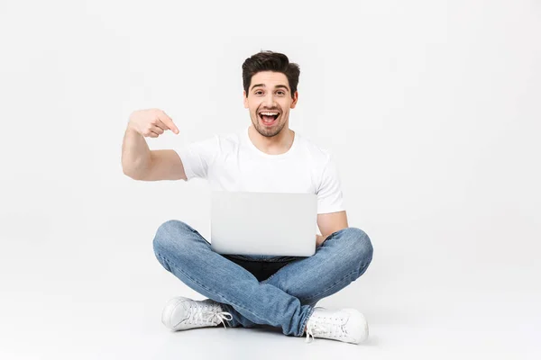 Heureux jeune homme excité posant isolé sur un mur blanc en utilisant un ordinateur portable pointant assis sur le sol . — Photo