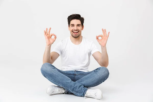 Aufgeregter emotionaler Mann posiert isoliert über weißem Wandhintergrund und zeigt okay Geste auf dem Boden sitzend. — Stockfoto