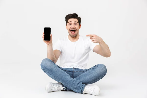 Emocionado joven posando aislado sobre fondo blanco de la pared utilizando el teléfono móvil que muestra la pantalla sentada en el suelo . — Foto de Stock