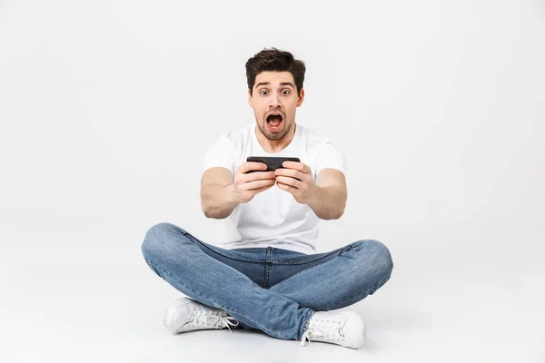 Shocked joven hombre posando aislado sobre fondo blanco de la pared jugar juegos por teléfono móvil . — Foto de Stock