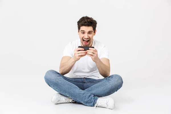 Feliz joven emocionado posando aislado sobre fondo de pared blanca jugar juegos por teléfono móvil . — Foto de Stock