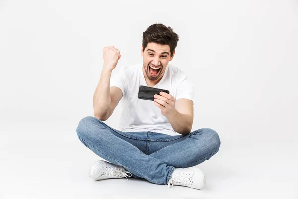 Ενθουσιασμένος νέος άνθρωπος που θέτει απομονωμένα πάνω από λευκό τοίχο παιχνίδι φόντο παιχνίδια από το κινητό τηλέφωνο κάνει νίκη χειρονομία. — Φωτογραφία Αρχείου