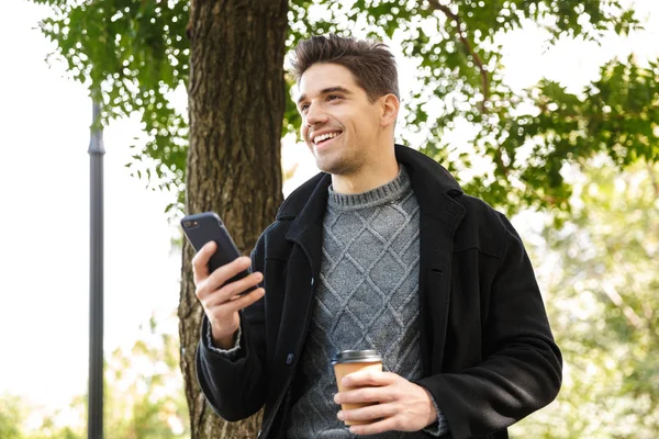 Handsome happy man in casual clothing walking outdoors in green park using mobile phone drinking coffee. — Stock Photo, Image